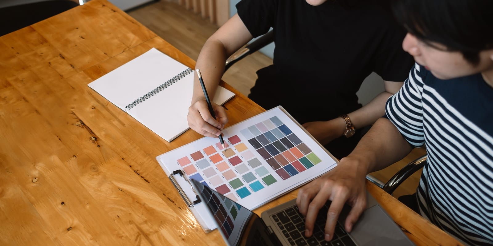 Two designers at a wooden table comparing printed colour swatches beside a laptop.