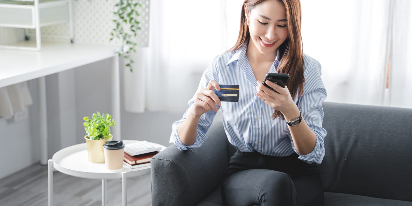 A woman on a sofa at home holds a bank card in one hand and a smartphone in the other.