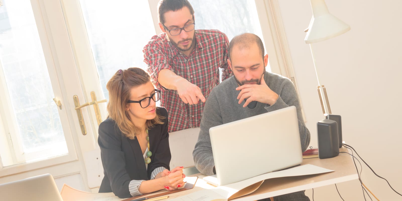 Three colleagues around a laptop at a sunlit desk, one pointing at the screen.