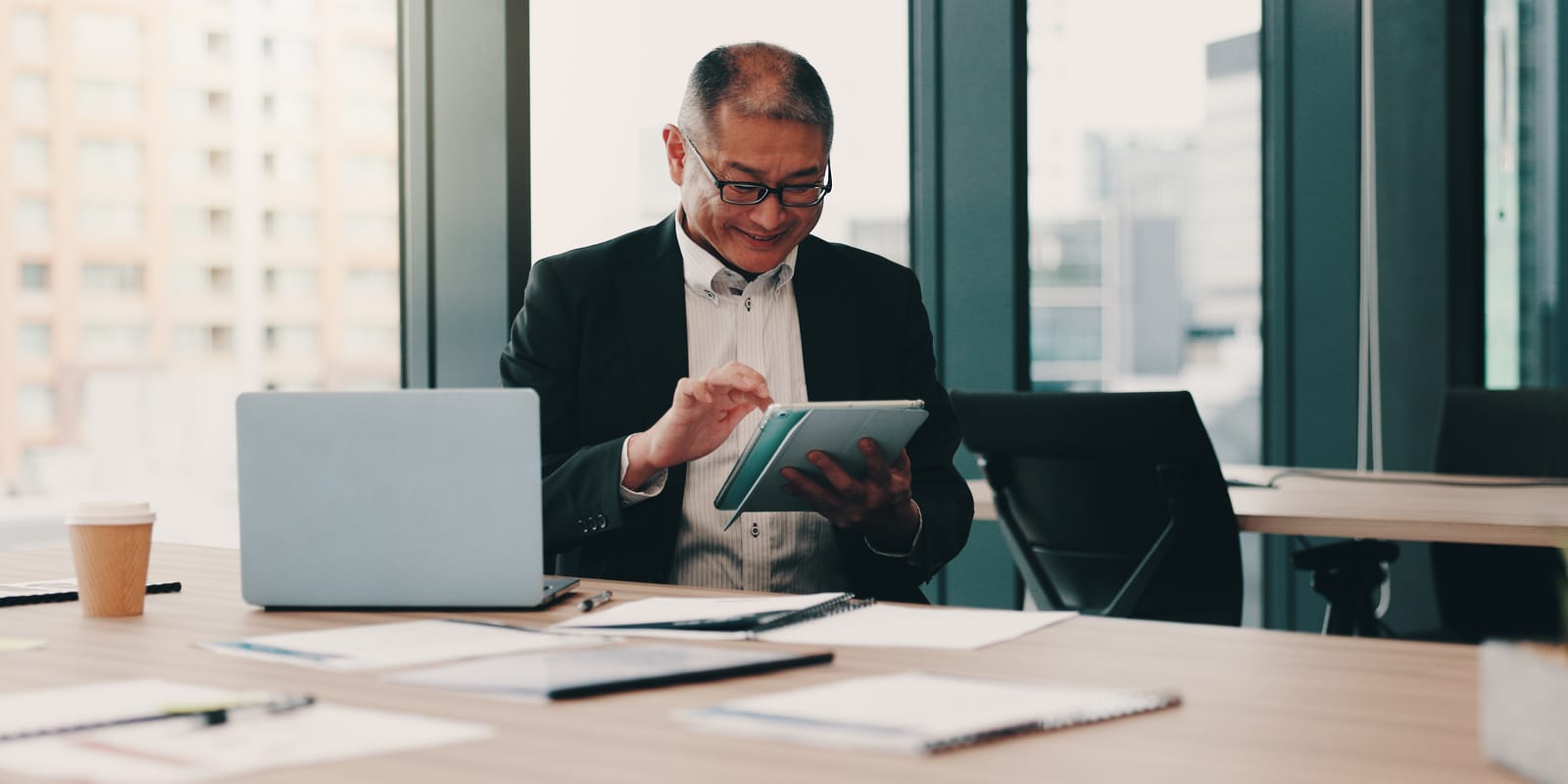 A man in a suit jacket reads from a tablet at an office desk, with a laptop and papers in front of him.