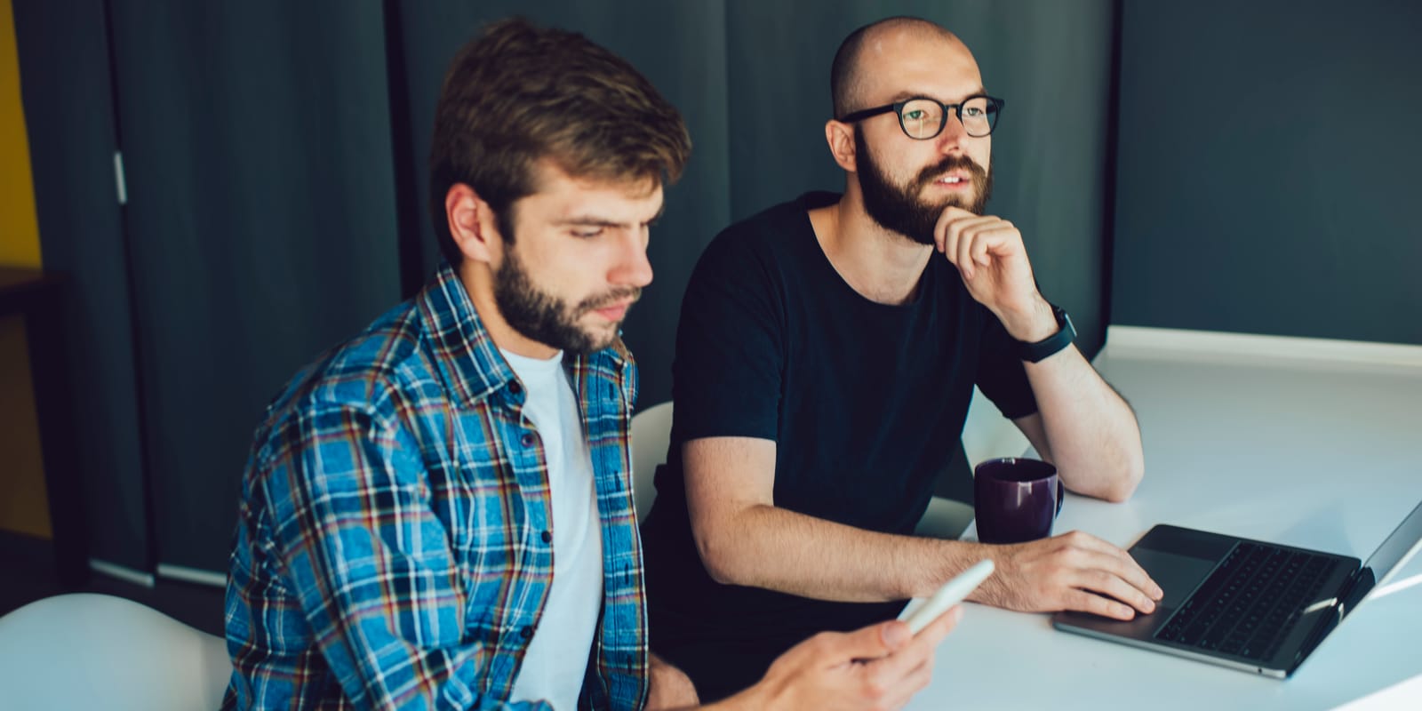 Two men at a desk look at a laptop together; one rests his hand at his chin.